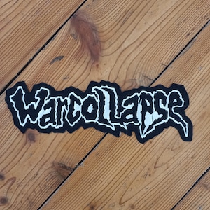 Può includere: Una toppa in bianco e nero con la scritta "Warcollapse" in un font stilizzato. La toppa ha un bordo nero ed è posta su uno sfondo di legno.