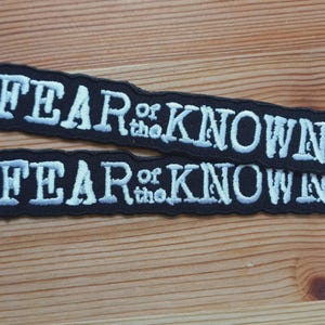 May include: Two black embroidered patches with the text "FEAR of the KNOWN" in white, stacked on top of each other. The patches have a curved, irregular shape and are set against a wooden background.