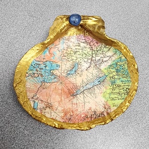 May include: A decorative trinket dish in the shape of a seashell. The interior displays a vintage map design, framed by a gold-painted edge. A small, blue, round stone adorns the top. Ideal for holding small items, such as jewellery.