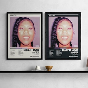Puede incluir: Dos carteles enmarcados con la portada del álbum "Her Loss" de Drake y 21 Savage. Los carteles muestran un retrato de una mujer con acentos rosas, el título del álbum y la lista de canciones. Los carteles están enmarcados en negro y montados en una estantería negra.