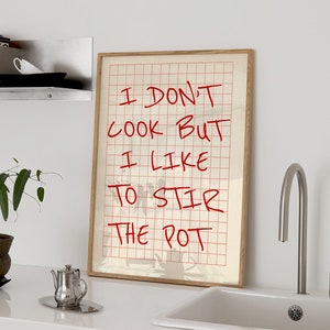 May include: A framed print with a red handwritten message on a gridded background. The message reads "I don't cook but I like to stir the pot". The print is hanging on a white wall above a white countertop with a stainless steel sink and faucet.