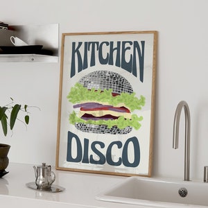 Pu&ograve; includere: Una stampa artistica incorniciata con un design a forma di hamburger. L'hamburger &egrave; composto da una palla da discoteca, lattuga, pomodoro e cipolla. Le parole "KITCHEN DISCO" sono scritte in blu scuro. La stampa ha una cornice in legno.