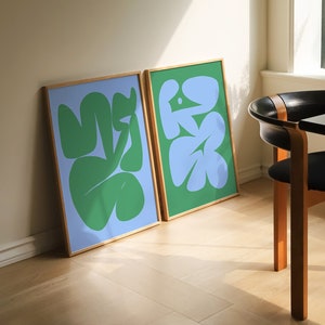 Op de afbeelding: Twee ingelijste abstracte kunstprints met groene en blauwe vormen op een lichtblauwe en groene achtergrond. De prints hangen aan een witte muur in een kamer met een houten vloer en een zwarte stoel.