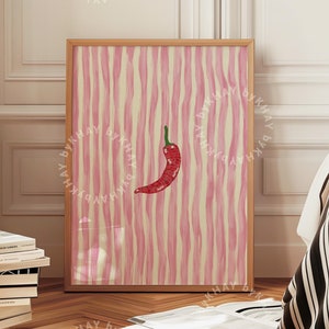 May include: A framed print of a red chili pepper on a pink and white striped background. The print is hanging on a white wall in a room with wooden floors.