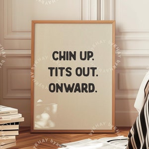 Op de afbeelding: Een ingelijst print met de tekst "Chin Up. Tits Out. Onward." op een beige achtergrond. De print hangt aan een witte muur in een kamer met een houten vloer en een gestreept deken op een bed.