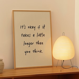 Puede incluir: Una impresión enmarcada con el texto "It's okay if it takes a little longer than you think." La impresión está en un estante de madera con una lámpara blanca y una pequeña planta en maceta.