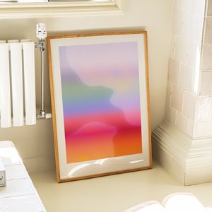 May include: A framed abstract print with a gradient of colours from purple to orange, hanging on a white wall next to a white radiator. A book and two paintbrushes are on the floor in front of the print.