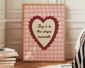 May include: A framed print with a pink gingham background and a heart-shaped design. The heart contains the text "Joy is in the simple moments" in a handwritten style. The frame is a light brown color. The print is leaning against a white wall.