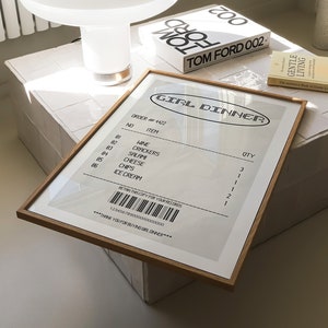 May include: A framed print with the text "GIRL DINNER" and a list of items, including wine, crackers, and ice cream. A white mushroom-shaped lamp and a book titled "TOM FORD 002" are also visible.