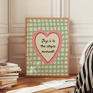 Könnte beinhalten: Ein gerahmter Druck mit dem Text "Joy is in the simple moments" in einer rosa Herzform. Der Hintergrund ist ein grün-weiß kariertes Muster. Der Rahmen ist hellbraun. Der Druck wird in einem Raum mit einem Bett und Büchern ausgestellt.