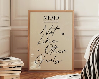 Puede incluir: Una impresión enmarcada con el texto "Not Like Other Girls" en una escritura elegante. La impresión tiene un fondo beige y un marco de madera. La imagen está en una habitación con una pila de libros y una cama con una manta a rayas.