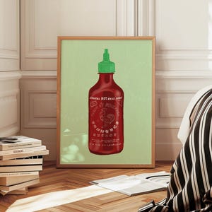 Op de afbeelding: Een ingelijste print met een rode Sriracha-chilisausfles met een groene dop. De fles heeft witte tekst en een hanenafbeelding, tegen een lichtgroene achtergrond. De lijst is lichtbruin.