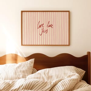 May include: A framed artwork with a pink and cream striped background and the handwritten text "Love like Jesus" in red. The artwork hangs above a bed with striped bedding and a wooden headboard.