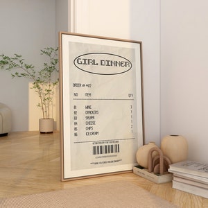 May include: A framed print with the words "GIRL DINNER" at the top. The print resembles a receipt, listing items like wine, crackers, salami, cheese, chips, and ice cream. The frame is a light brown color.