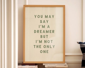 May include: A framed print with a white background and green text that reads "You may say I'm a dreamer but I'm not the only one". The print is sitting on a stack of books.