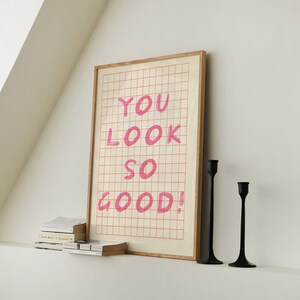 K&ouml;nnte beinhalten: Ein gerahmter Druck mit einem Gittermuster und dem Text "YOU LOOK SO GOOD!" in rosa. Zwei schwarze Kerzenhalter stehen auf einer wei&szlig;en Oberfl&auml;che unter dem Druck.