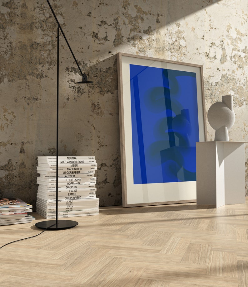 May include: A stack of books with the names of famous architects on the spines, a black floor lamp with a white shade, a white pedestal with a white abstract sculpture, and a framed blue abstract painting on a white wall.