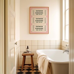 May include: A white bathroom with a white bathtub, a wooden stool, and a framed print of three pink tickets that say "Shower Karaoke One Entry".