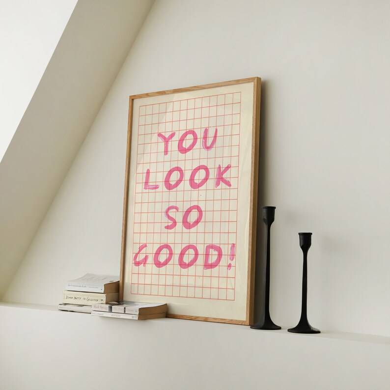 K&ouml;nnte beinhalten: Ein gerahmter Druck mit einem Gittermuster und dem Text "YOU LOOK SO GOOD!" in rosa. Zwei schwarze Kerzenhalter stehen auf einer wei&szlig;en Oberfl&auml;che unter dem Druck.