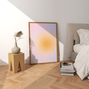 May include: A wooden side table with a vase of dried flowers and a framed abstract print with a gradient of pink, purple, and orange colors. The print has a light brown frame and is hanging on a white wall.