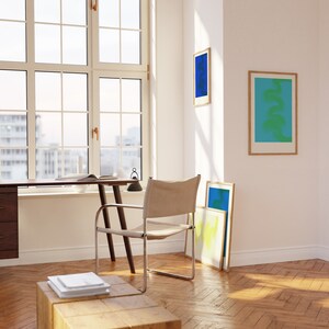 May include: A wooden desk with a chair and a stack of books on a wooden stool. There are three framed abstract art prints on the wall behind the desk. The room has a large window with a view of a city.
