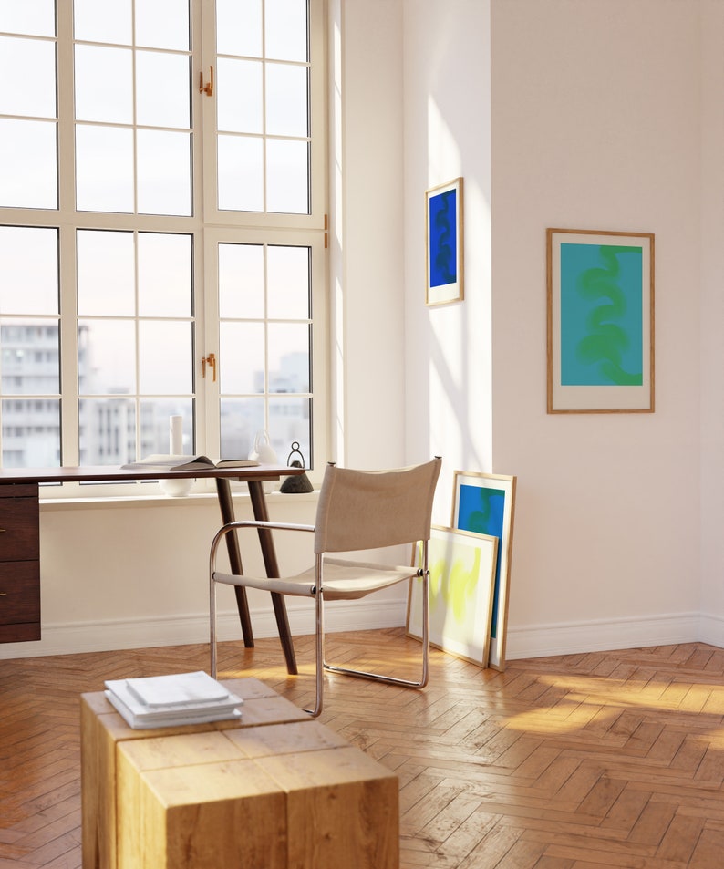 May include: A wooden desk with a chair and a stack of books on a wooden stool. There are three framed abstract art prints on the wall behind the desk. The room has a large window with a view of a city.