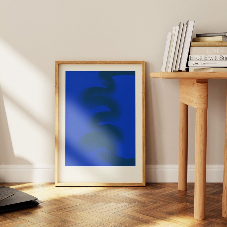 May include: A framed abstract art print with a blue background and a dark blue abstract shape in the center. The frame is made of wood and has a light brown finish.