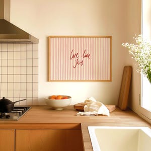 May include: A kitchen scene with a wooden countertop, a white sink, and a framed artwork. The artwork features pink and white vertical stripes with the handwritten text "love like Jesus". A bowl of oranges sits on the counter.