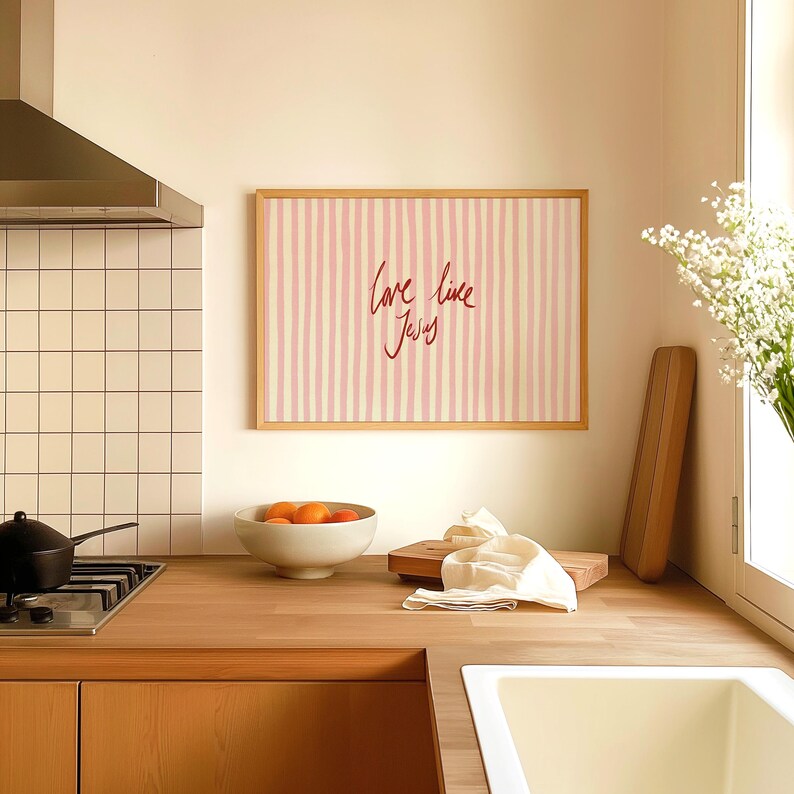 May include: A kitchen scene with a wooden countertop, a white sink, and a framed artwork. The artwork features pink and white vertical stripes with the handwritten text "love like Jesus". A bowl of oranges sits on the counter.