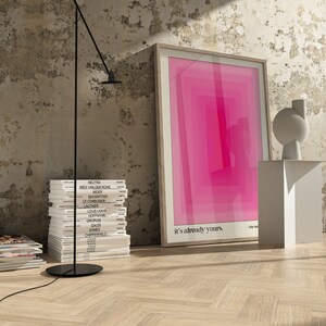May include: A framed pink and white abstract print with the text "it's already yours." hanging on a white wall with a stack of books and a black lamp.