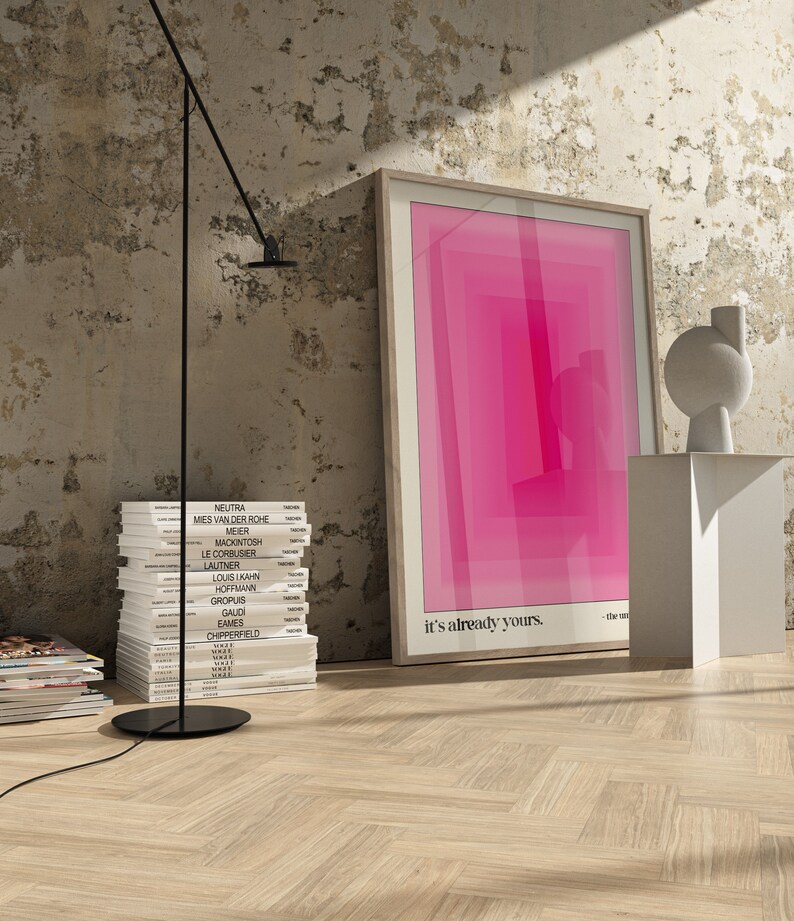 May include: A framed pink and white abstract print with the text "it's already yours." hanging on a white wall with a stack of books and a black lamp.