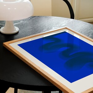May include: A framed abstract art print with a blue background and dark blue abstract shapes. The print is sitting on a black table.