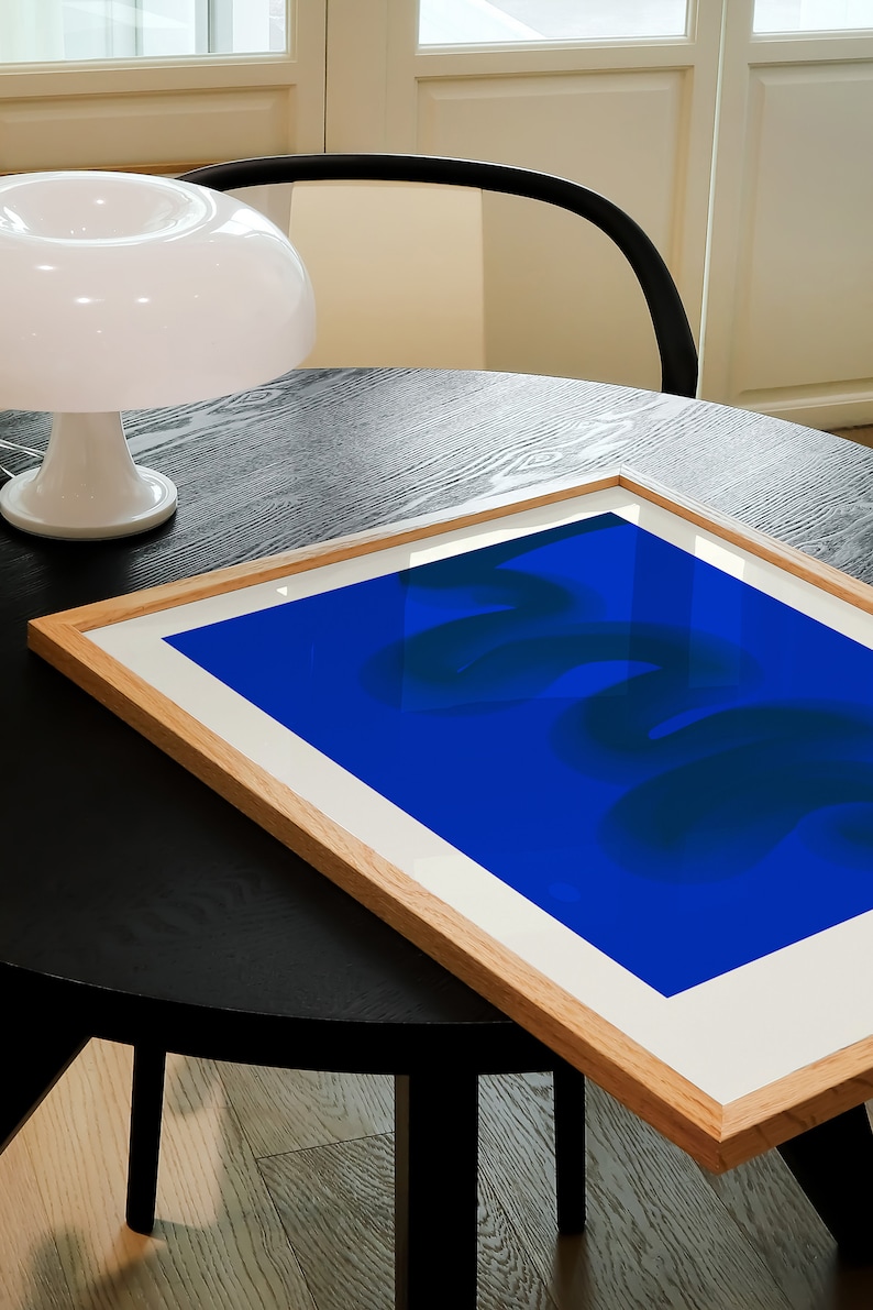 May include: A framed abstract art print with a blue background and dark blue abstract shapes. The print is sitting on a black table.