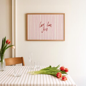 May include: A framed artwork with a light wood frame hangs on a white wall. The artwork features pink and white vertical stripes with the words "love like Jesus" in red script. A table is set with a striped tablecloth, glasses, and tulips.