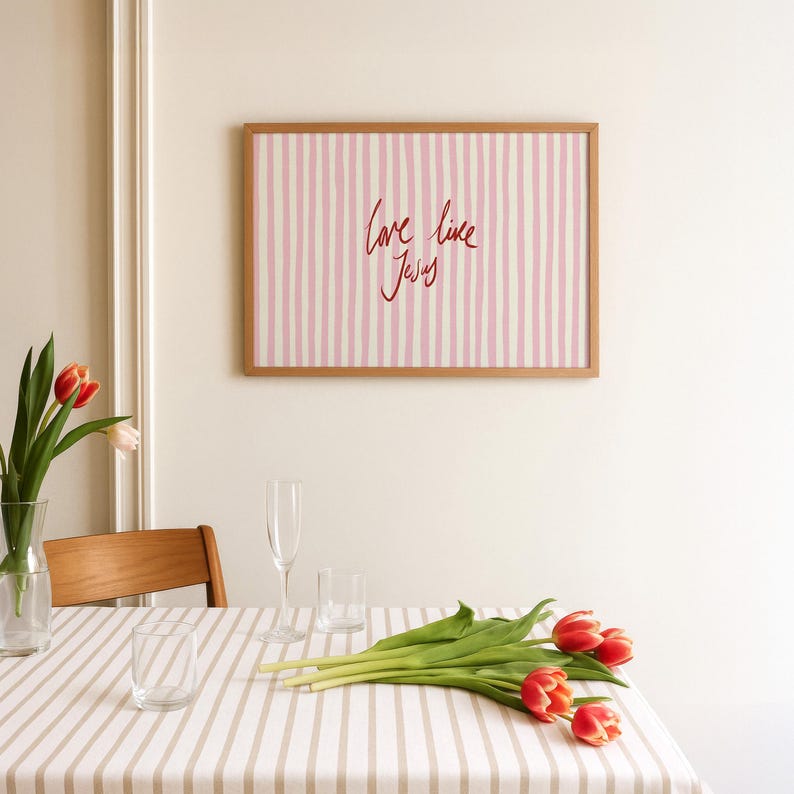 May include: A framed artwork with a light wood frame hangs on a white wall. The artwork features pink and white vertical stripes with the words "love like Jesus" in red script. A table is set with a striped tablecloth, glasses, and tulips.