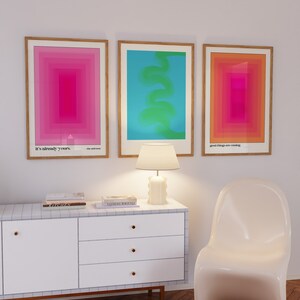 May include: Three framed abstract art prints with geometric shapes and gradients in pink, blue, and orange. The prints are hung on a white wall above a white dresser with three drawers and gold handles. A white lamp with a white shade sits on the dresser. A white chair sits in front of the dresser.