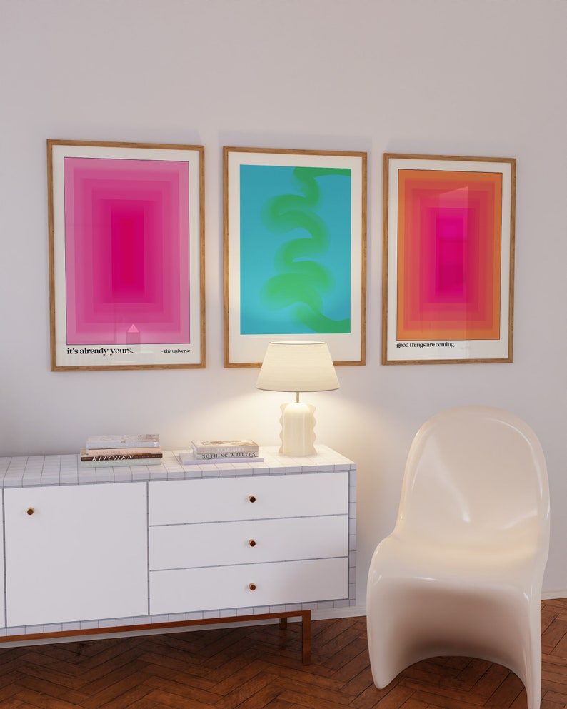 May include: Three framed abstract art prints with geometric shapes and gradients in pink, blue, and orange. The prints are hung on a white wall above a white dresser with three drawers and gold handles. A white lamp with a white shade sits on the dresser. A white chair sits in front of the dresser.