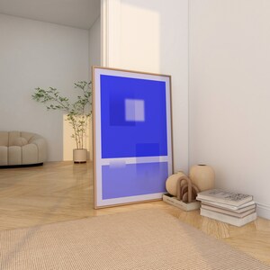 May include: A framed abstract art print with a blue background and white geometric shapes. The print is leaning against a white wall next to a stack of books and decorative vases.