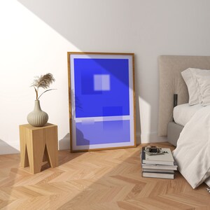 May include: A wooden side table with a vase of dried flowers and a framed abstract painting with a blue and white color scheme. The painting is hanging on a white wall above the table. A bed with white linens is in the background.