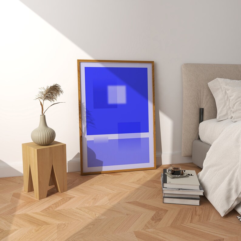 May include: A wooden side table with a vase of dried flowers and a framed abstract painting with a blue and white color scheme. The painting is hanging on a white wall above the table. A bed with white linens is in the background.