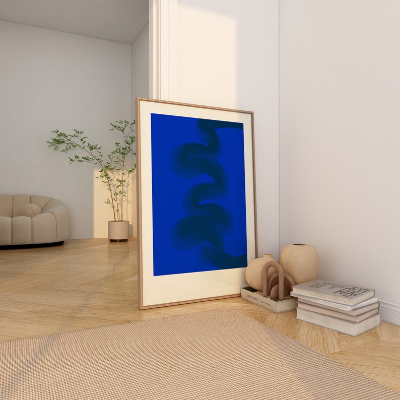 May include: A framed abstract art print with a blue background and a dark blue abstract design. The print is leaning against a white wall next to a small wooden table with a stack of books and decorative vases.