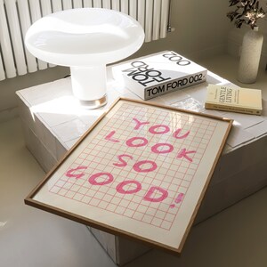 K&ouml;nnte beinhalten: Eine wei&szlig;e Keramik-Tischlampe mit einem pilzf&ouml;rmigen Schirm steht auf einer wei&szlig;en, gefliesten Oberfl&auml;che. Ein gerahmter Druck mit dem Text "YOU LOOK SO GOOD!" in rosa Schrift befindet sich auf dem Tisch. Zwei B&uuml;cher, "TOM FORD 002" und "The Japanese Book of Gentle Living", liegen ebenfalls auf dem Tisch.