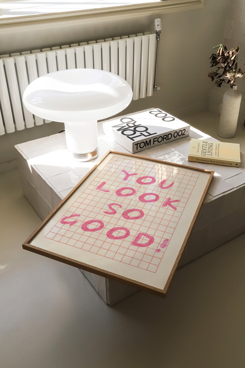 K&ouml;nnte beinhalten: Eine wei&szlig;e Keramik-Tischlampe mit einem pilzf&ouml;rmigen Schirm steht auf einer wei&szlig;en, gefliesten Oberfl&auml;che. Ein gerahmter Druck mit dem Text "YOU LOOK SO GOOD!" in rosa Schrift befindet sich auf dem Tisch. Zwei B&uuml;cher, "TOM FORD 002" und "The Japanese Book of Gentle Living", liegen ebenfalls auf dem Tisch.