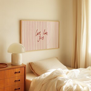 May include: A bedroom scene featuring a framed artwork with pink and white vertical stripes and the text "Love like Jesus" in red script. A white mushroom-shaped lamp sits on a wooden dresser. The bed is made with cream-colored linens.