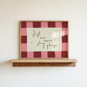 Peut inclure: Une affiche encadrée avec l'inscription manuscrite "Just one more story please". L'affiche a une bordure à carreaux rose et bordeaux et est exposée sur une étagère en bois.