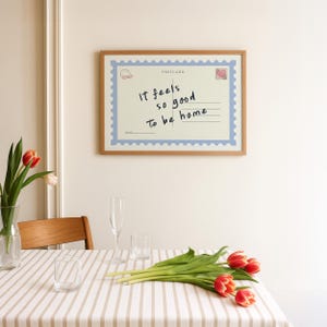 May include: A framed postcard print with the text "It feels so good to be home." The print has a light blue border and a postage stamp design. The frame is made of wood. A table is set with a striped tablecloth, glasses, and tulips.