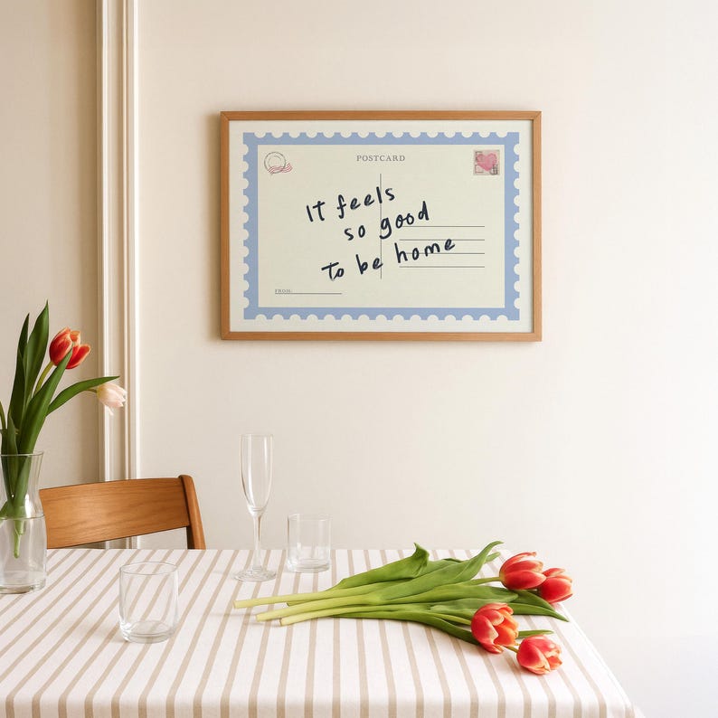 May include: A framed postcard print with the text "It feels so good to be home." The print has a light blue border and a postage stamp design. The frame is made of wood. A table is set with a striped tablecloth, glasses, and tulips.
