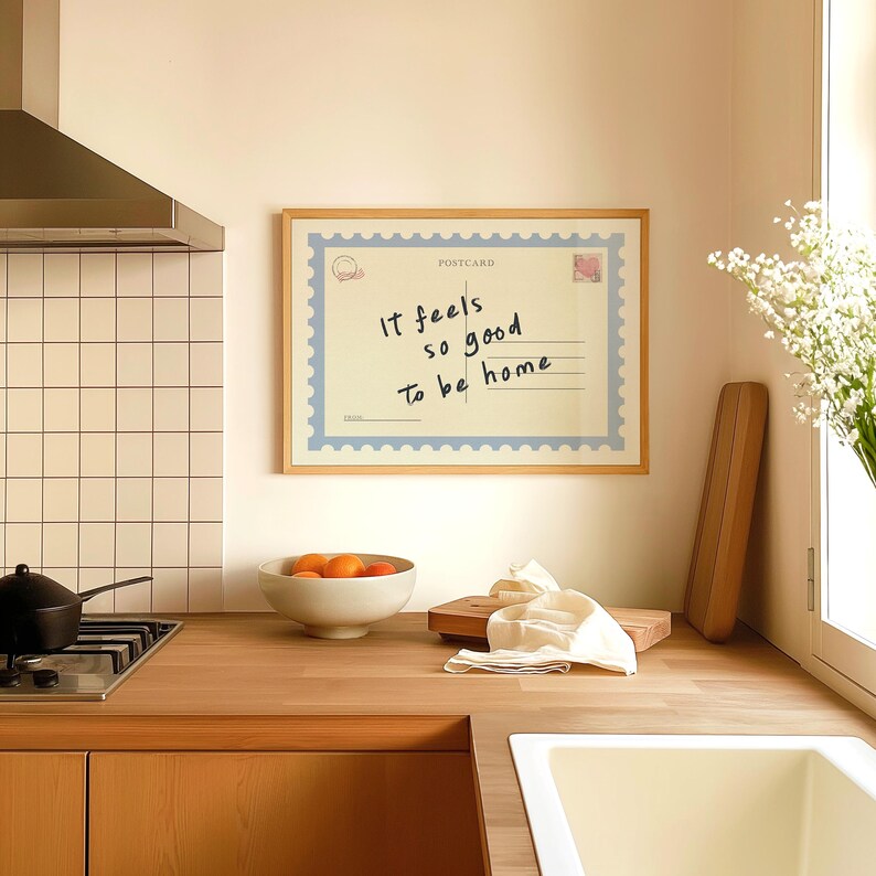 May include: A framed postcard with the text "It feels so good to be home" hangs on a wall in a kitchen. The postcard is light yellow with a blue border and a postage stamp design. A bowl of oranges, a cutting board, and a white cloth are on the wooden countertop.