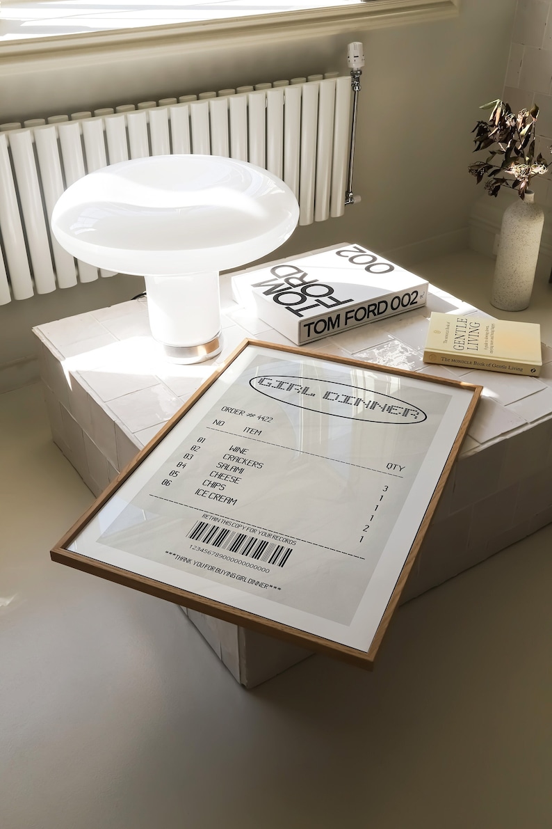 May include: A framed print with the text "GIRL DINNER" and a list of items, including wine, crackers, and ice cream. A white mushroom-shaped lamp and a book titled "TOM FORD 002" are also visible.