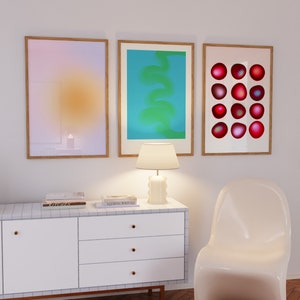 May include: Three framed abstract art prints on a white wall above a white dresser with three drawers. The prints are in a light pink, blue, and white color scheme. The dresser has a white top and gold knobs. A white lamp sits on the dresser. A white chair is in the foreground.